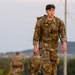 A U.S. Army soldier in Operational Camouflage Pattern (OCP) uniform is conducting a ruck march, carrying a loaded rucksack. The soldier is in the foreground and is clearly identifiable by 'U.S. ARMY' chest tape and an American flag patch on the left shoulder. Additional soldiers in similar uniforms are visible in the blurred background, suggesting a group training exercise.