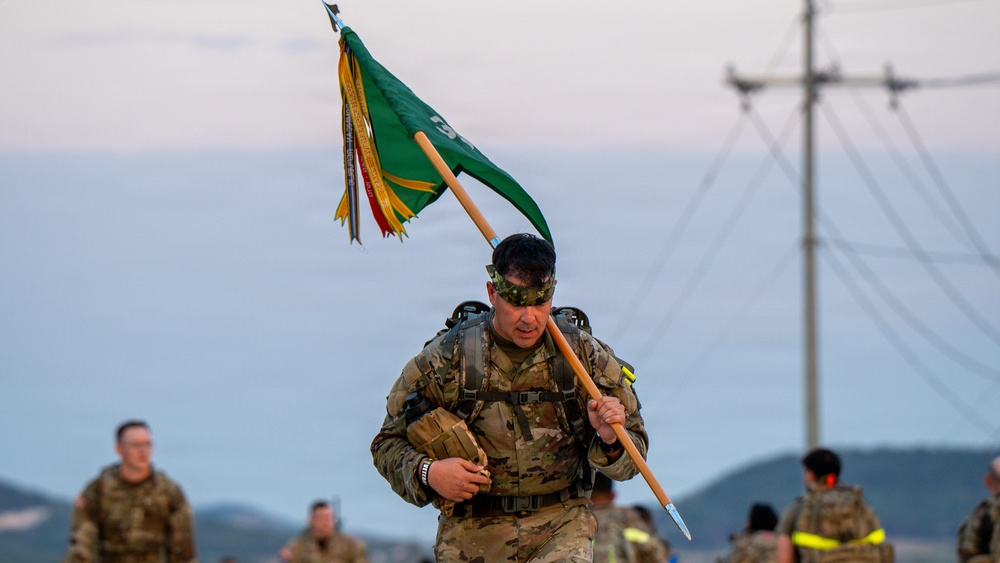 A soldier in full combat uniform and gear carries a military unit guidon (green flag) while marching or rucking, likely during a military training exercise or ruck march event. The soldier wears camouflage face paint and carries a heavy rucksack, suggesting a physically demanding field exercise. Additional soldiers in similar gear are visible in the background against a coastal or hilly landscape.