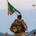 A soldier in full combat uniform and gear carries a military unit guidon (green flag) while marching or rucking, likely during a military training exercise or ruck march event. The soldier wears camouflage face paint and carries a heavy rucksack, suggesting a physically demanding field exercise. Additional soldiers in similar gear are visible in the background against a coastal or hilly landscape.