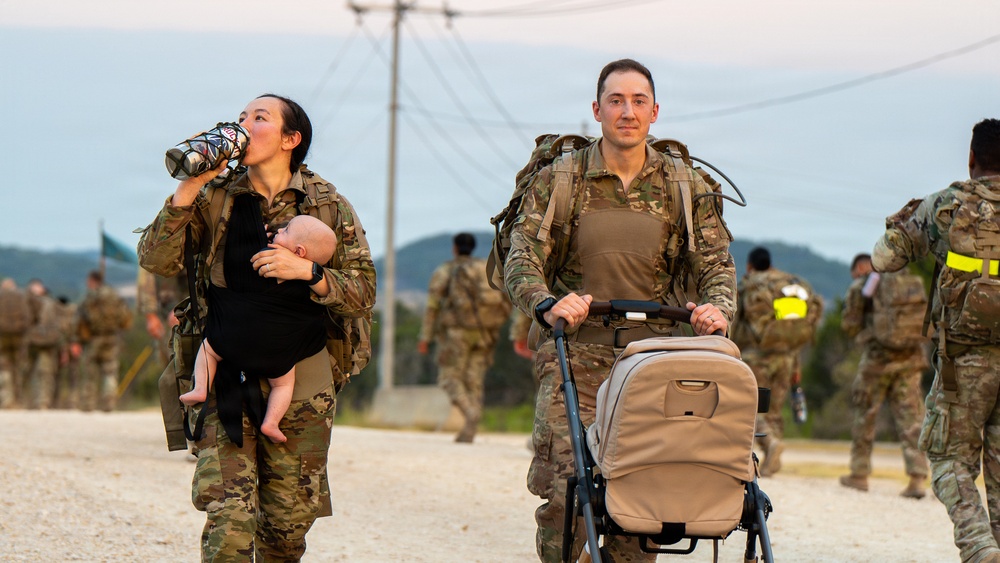 Two soldiers in camouflage uniforms and tactical gear are participating in what appears to be a military ruck march or physical training event. A female soldier carries an infant while drinking from a water bottle, and a male soldier pushes an empty stroller alongside her. Additional uniformed personnel are visible in the background on a gravel road with hills and utility poles.