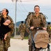 Two soldiers in camouflage uniforms and tactical gear are participating in what appears to be a military ruck march or physical training event. A female soldier carries an infant while drinking from a water bottle, and a male soldier pushes an empty stroller alongside her. Additional uniformed personnel are visible in the background on a gravel road with hills and utility poles.
