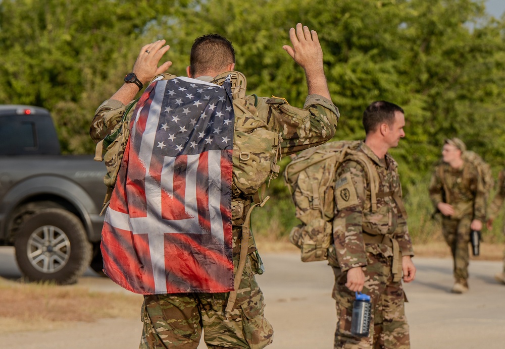 The image shows military personnel in multicam/OCP camouflage uniforms carrying tactical rucksacks, likely during a ruck march or physical training event. The foreground soldier has an American flag draped over their back and has both hands raised above their head, a common posture during strenuous physical activity. At least three soldiers are visible, with additional personnel blurred in the background.