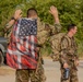The image shows military personnel in multicam/OCP camouflage uniforms carrying tactical rucksacks, likely during a ruck march or physical training event. The foreground soldier has an American flag draped over their back and has both hands raised above their head, a common posture during strenuous physical activity. At least three soldiers are visible, with additional personnel blurred in the background.