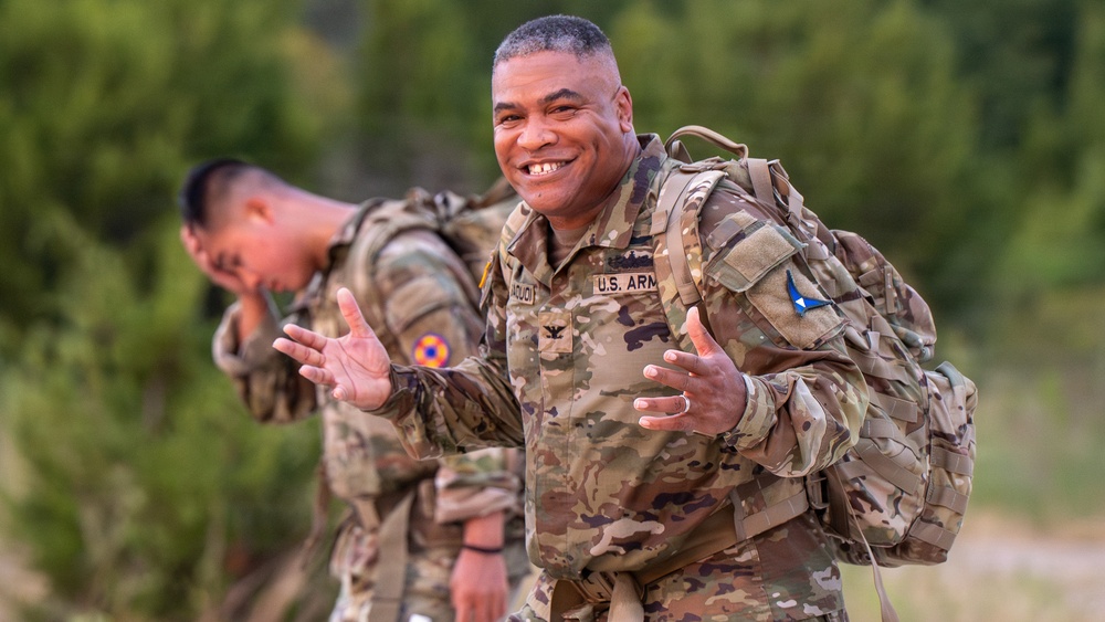 A U.S. Army soldier in the foreground smiles broadly while carrying a military rucksack in an outdoor training environment. He is wearing OCP (Operational Camouflage Pattern) uniform with visible rank insignia of a Colonel (eagle) and a name tape partially reading 'AGUCI' or similar. A second soldier is visible in the background, also in full gear and appearing to be exerting effort.