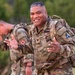 A U.S. Army soldier in the foreground smiles broadly while carrying a military rucksack in an outdoor training environment. He is wearing OCP (Operational Camouflage Pattern) uniform with visible rank insignia of a Colonel (eagle) and a name tape partially reading 'AGUCI' or similar. A second soldier is visible in the background, also in full gear and appearing to be exerting effort.