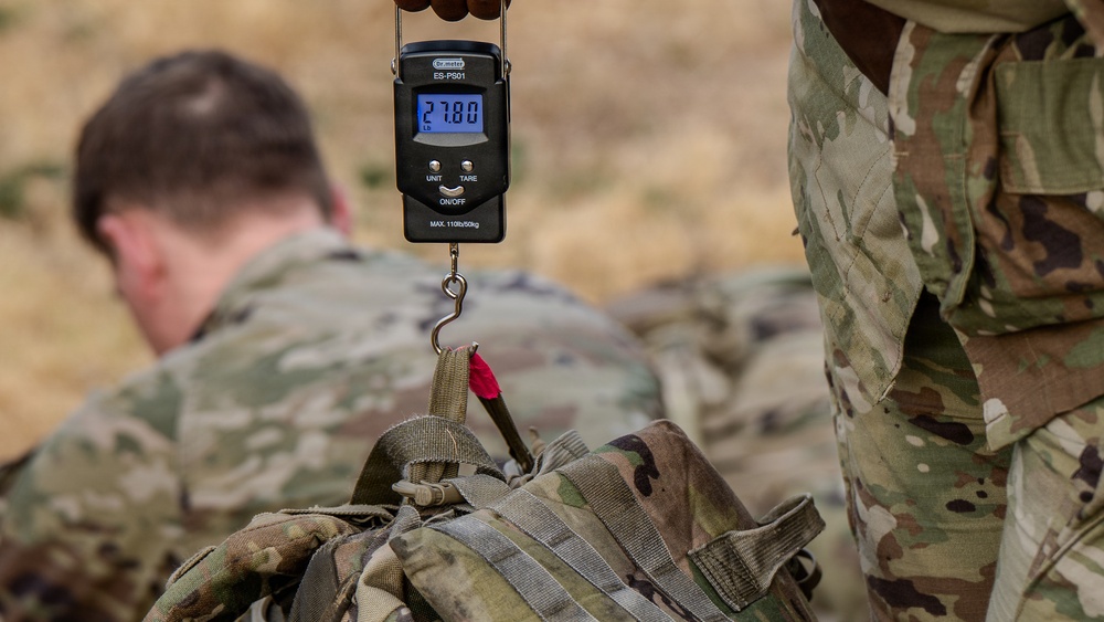 A military personnel is using a digital hanging scale (model ES-PS01) to weigh a camouflage tactical backpack, with the scale displaying 27.80 lbs. A second soldier in MultiCam/OCP camouflage uniform is visible in the blurred background, crouching or kneeling on arid terrain. This appears to be a gear inspection or load verification activity in a field training or operational environment.