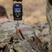 A military personnel is using a digital hanging scale (model ES-PS01) to weigh a camouflage tactical backpack, with the scale displaying 27.80 lbs. A second soldier in MultiCam/OCP camouflage uniform is visible in the blurred background, crouching or kneeling on arid terrain. This appears to be a gear inspection or load verification activity in a field training or operational environment.