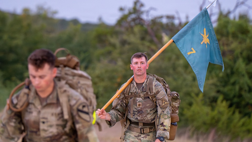 Two soldiers in U.S. Army multicam camouflage uniforms are conducting a ruck march while carrying heavy backpacks. The soldier in the foreground carries a light blue unit guidon flag with a gold insignia and the number '7' visible. The setting appears to be a rural or training area with trees and overcast skies in the background.