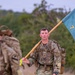 Two soldiers in U.S. Army multicam camouflage uniforms are conducting a ruck march while carrying heavy backpacks. The soldier in the foreground carries a light blue unit guidon flag with a gold insignia and the number '7' visible. The setting appears to be a rural or training area with trees and overcast skies in the background.