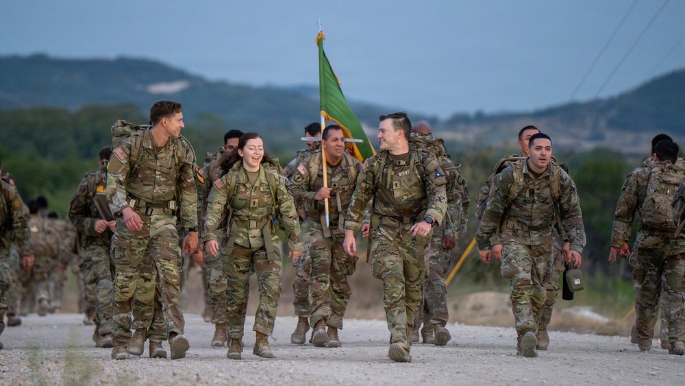 A group of U.S. Army soldiers in MultiCam/OCP camouflage uniforms are conducting a ruck march along a gravel road in what appears to be a training environment. The soldiers are carrying rucksacks and one is bearing a green unit guidon flag. The mood appears positive, with several soldiers smiling and laughing during the march.