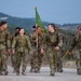 A group of U.S. Army soldiers in MultiCam/OCP camouflage uniforms are conducting a ruck march along a gravel road in what appears to be a training environment. The soldiers are carrying rucksacks and one is bearing a green unit guidon flag. The mood appears positive, with several soldiers smiling and laughing during the march.