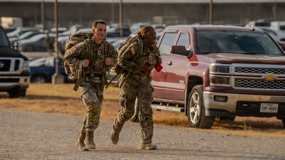 Two U.S. Army soldiers in MultiCam (OCP) camouflage uniforms are running while carrying large rucksacks in what appears to be a military installation parking lot. Both soldiers wear American flag patches on their shoulders and carry full field gear, suggesting a ruck march or physical training event. The setting appears to be in Texas, based on a visible license plate on a nearby red Chevrolet pickup truck.