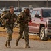 Two U.S. Army soldiers in MultiCam (OCP) camouflage uniforms are running while carrying large rucksacks in what appears to be a military installation parking lot. Both soldiers wear American flag patches on their shoulders and carry full field gear, suggesting a ruck march or physical training event. The setting appears to be in Texas, based on a visible license plate on a nearby red Chevrolet pickup truck.