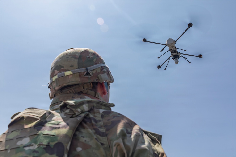 A military personnel wearing camouflage uniform and helmet is observing a multi-rotor drone (hexacopter or octocopter) in flight against a clear blue sky. The drone appears to be a large tactical unmanned aerial vehicle equipped with a camera or sensor payload suspended beneath its frame. The image is taken from a low angle behind the soldier, emphasizing the drone in the upper right portion of the frame.