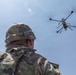 A military personnel wearing camouflage uniform and helmet is observing a multi-rotor drone (hexacopter or octocopter) in flight against a clear blue sky. The drone appears to be a large tactical unmanned aerial vehicle equipped with a camera or sensor payload suspended beneath its frame. The image is taken from a low angle behind the soldier, emphasizing the drone in the upper right portion of the frame.