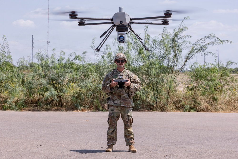 A military service member in full camouflage uniform is operating a large multi-rotor drone (hexacopter or octocopter) using a handheld remote controller. The drone is hovering directly above the operator, equipped with what appears to be a camera or sensor payload underneath. The scene takes place on a paved surface in an arid, semi-vegetated outdoor environment.