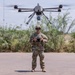 A military service member in full camouflage uniform is operating a large multi-rotor drone (hexacopter or octocopter) using a handheld remote controller. The drone is hovering directly above the operator, equipped with what appears to be a camera or sensor payload underneath. The scene takes place on a paved surface in an arid, semi-vegetated outdoor environment.