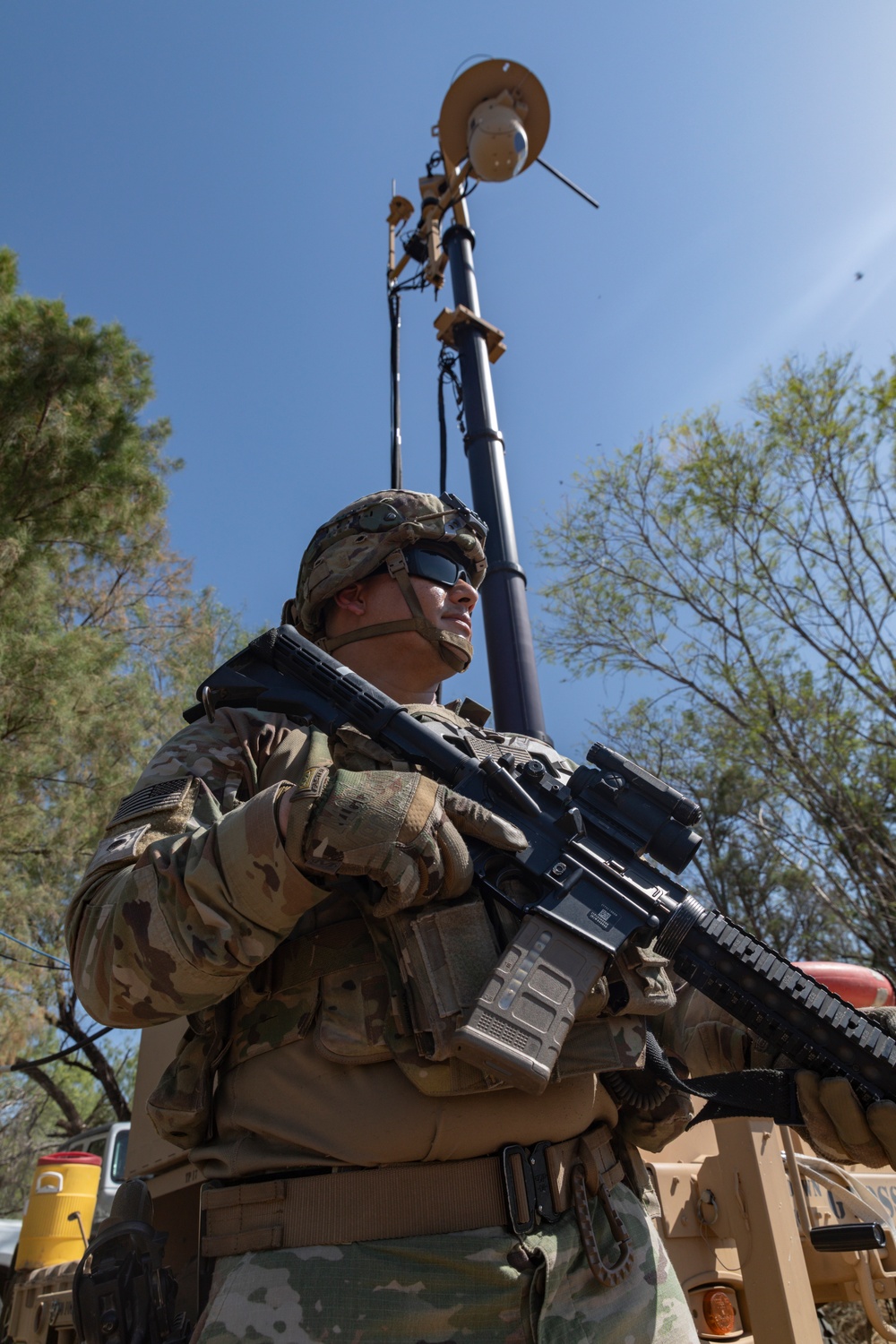 A U.S. Army soldier in full combat gear stands at alert while holding an M4-style carbine rifle. Behind the soldier, a telescoping surveillance or communications mast is deployed, likely mounted on a military vehicle. The setting appears to be an outdoor area with trees and clear blue sky, suggesting a field operation or border security mission.