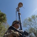 A U.S. Army soldier in full combat gear stands at alert while holding an M4-style carbine rifle. Behind the soldier, a telescoping surveillance or communications mast is deployed, likely mounted on a military vehicle. The setting appears to be an outdoor area with trees and clear blue sky, suggesting a field operation or border security mission.