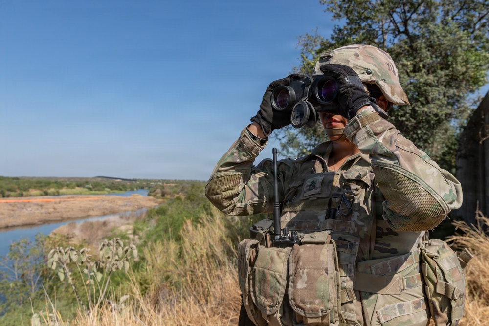A soldier in full combat gear is using binoculars to observe a landscape featuring a river and open terrain. The soldier is wearing OCP (Operational Camouflage Pattern) uniform, tactical vest, helmet, and black gloves, consistent with U.S. Army equipment. The setting appears to be an elevated observation point overlooking a river valley under clear blue skies.
