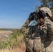 A soldier in full combat gear is using binoculars to observe a landscape featuring a river and open terrain. The soldier is wearing OCP (Operational Camouflage Pattern) uniform, tactical vest, helmet, and black gloves, consistent with U.S. Army equipment. The setting appears to be an elevated observation point overlooking a river valley under clear blue skies.