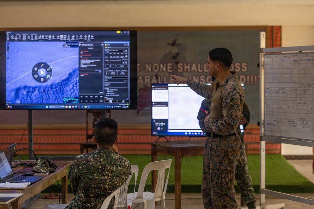 Two military personnel in camouflage uniforms are working in what appears to be a tactical operations center or command post. One soldier is seated at a desk with a laptop while another stands and points at a large display screen showing what appears to be drone or aerial surveillance footage with a targeting interface. A whiteboard with a grid/tracking chart is visible to the right.