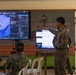 Two military personnel in camouflage uniforms are working in what appears to be a tactical operations center or command post. One soldier is seated at a desk with a laptop while another stands and points at a large display screen showing what appears to be drone or aerial surveillance footage with a targeting interface. A whiteboard with a grid/tracking chart is visible to the right.