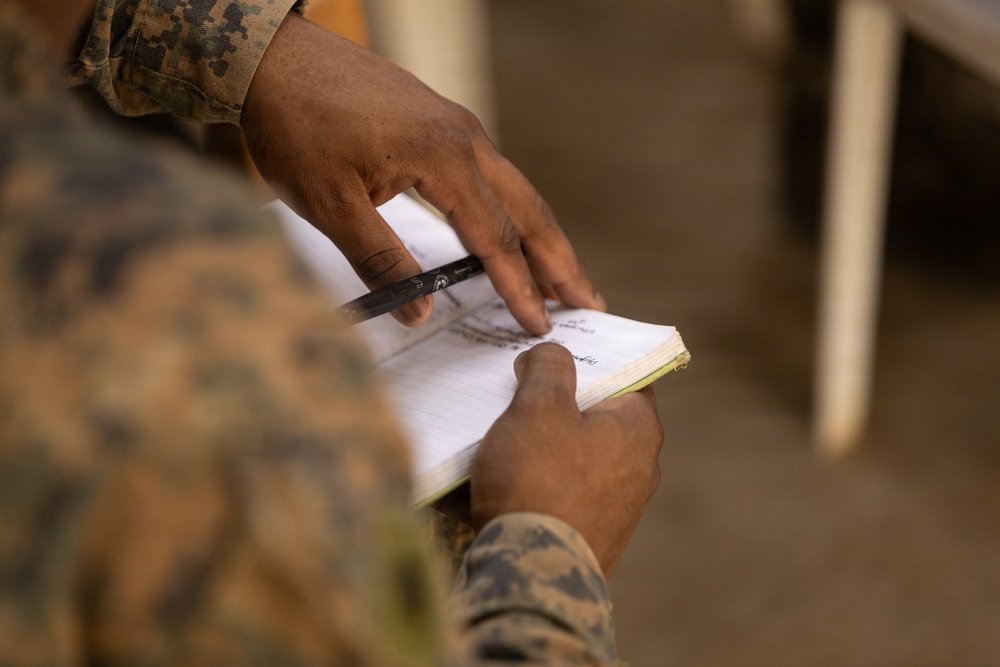 A close-up photograph of a person in military camouflage uniform holding a small notebook and a pen. The individual appears to be reviewing or taking notes in the notebook. The background is blurred, suggesting an outdoor or semi-outdoor environment.