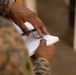A close-up photograph of a person in military camouflage uniform holding a small notebook and a pen. The individual appears to be reviewing or taking notes in the notebook. The background is blurred, suggesting an outdoor or semi-outdoor environment.