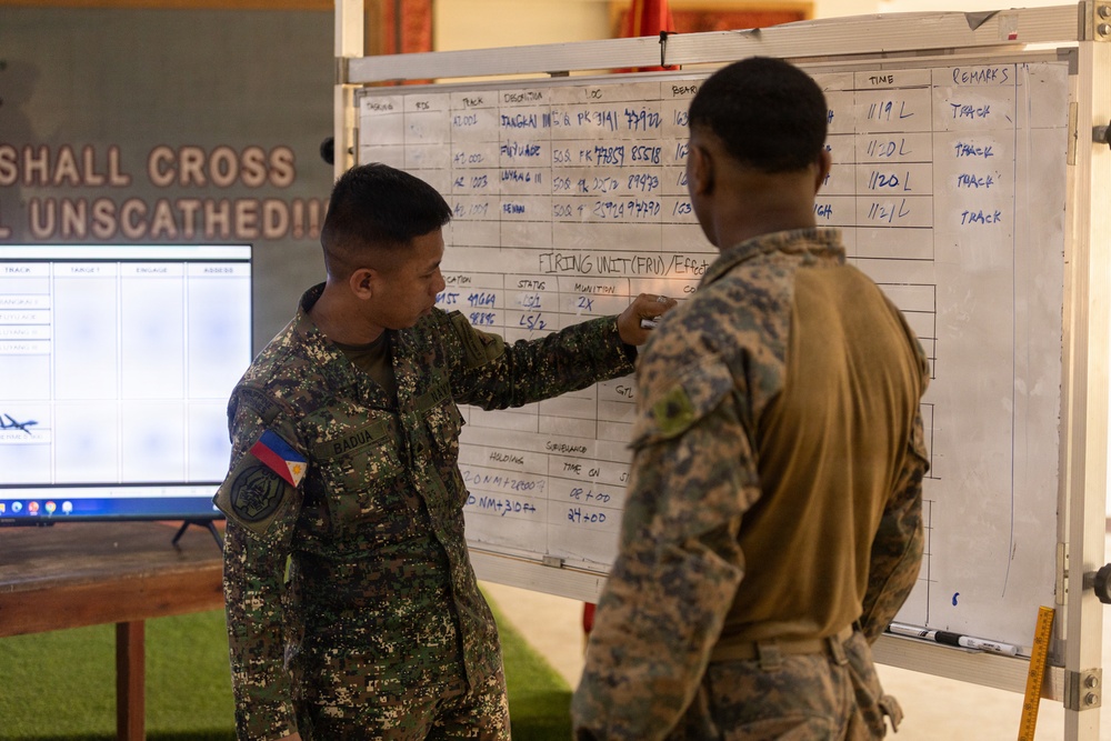 Two military personnel in camouflage uniforms are reviewing a whiteboard containing tactical information including target tracking data, firing unit details, and time logs. One soldier, identified by a Philippine flag patch on his shoulder with the name 'BADUA' on his uniform, is pointing at the whiteboard while the other soldier observes. The setting appears to be a military operations or briefing room.