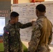 Two military personnel in camouflage uniforms are reviewing a whiteboard containing tactical information including target tracking data, firing unit details, and time logs. One soldier, identified by a Philippine flag patch on his shoulder with the name 'BADUA' on his uniform, is pointing at the whiteboard while the other soldier observes. The setting appears to be a military operations or briefing room.
