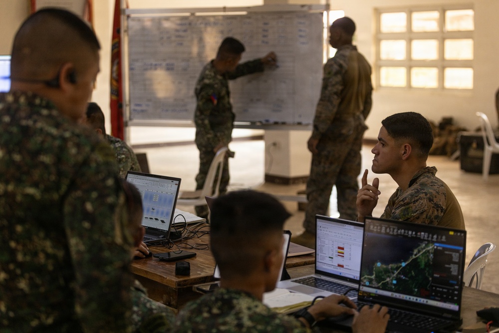 Military personnel from what appear to be two different armed forces are engaged in a joint planning or briefing session inside a building. Soldiers are working at laptops displaying maps and data while one individual writes on a whiteboard at the front. The setting suggests a tactical operations or coordination exercise.