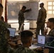 Military personnel from what appear to be two different armed forces are engaged in a joint planning or briefing session inside a building. Soldiers are working at laptops displaying maps and data while one individual writes on a whiteboard at the front. The setting suggests a tactical operations or coordination exercise.
