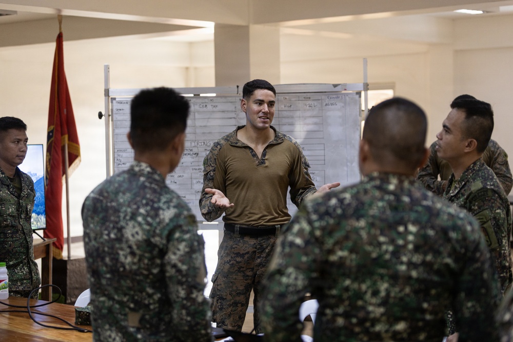 A military briefing or training session is taking place indoors, with one individual in a solid olive/tan combat shirt addressing a group of soldiers wearing digital camouflage uniforms. A whiteboard with written content is visible in the background, along with what appears to be a military unit flag or guidon. The setting suggests a joint military exercise or instructional session.