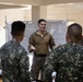 A military briefing or training session is taking place indoors, with one individual in a solid olive/tan combat shirt addressing a group of soldiers wearing digital camouflage uniforms. A whiteboard with written content is visible in the background, along with what appears to be a military unit flag or guidon. The setting suggests a joint military exercise or instructional session.