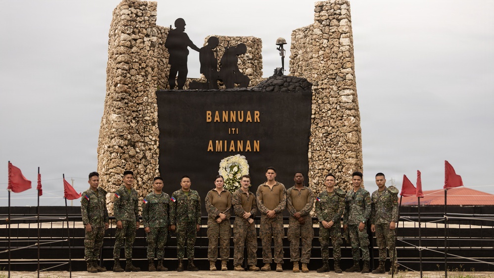 A group of military personnel pose in front of a memorial monument inscribed with 'BANNUAR ITI AMIANAN,' which translates from Ilocano (Filipino dialect) to 'Hero of the North.' The monument features stone pillars, silhouetted soldier figures, a battlefield cross, and a commemorative wreath, suggesting a joint military ceremony. The group includes both Philippine Armed Forces soldiers and what appear to be U.S. Marines in different camouflage uniforms.