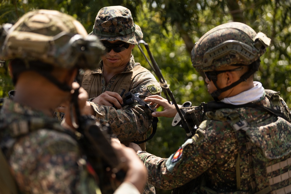 The image shows military personnel from what appears to be two different armed forces engaged in a joint training or equipment check activity in an outdoor wooded setting. A central figure wearing a MARPAT (US Marine Corps) camouflage pattern and boonie hat is interacting with soldiers wearing a different camouflage pattern and tactical helmets. The interaction appears cooperative and instructional in nature.