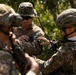 The image shows military personnel from what appears to be two different armed forces engaged in a joint training or equipment check activity in an outdoor wooded setting. A central figure wearing a MARPAT (US Marine Corps) camouflage pattern and boonie hat is interacting with soldiers wearing a different camouflage pattern and tactical helmets. The interaction appears cooperative and instructional in nature.