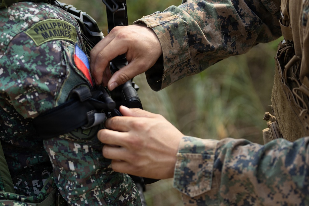 Two military personnel are shown in close interaction, with one adjusting or inspecting equipment on the other. The soldier on the left wears a 'Philippine Marines' patch with a Philippine flag insignia, while the soldier on the right wears USMC MARPAT digital camouflage. This appears to depict a joint training or cooperative military exercise between Philippine Marines and U.S. Marines.