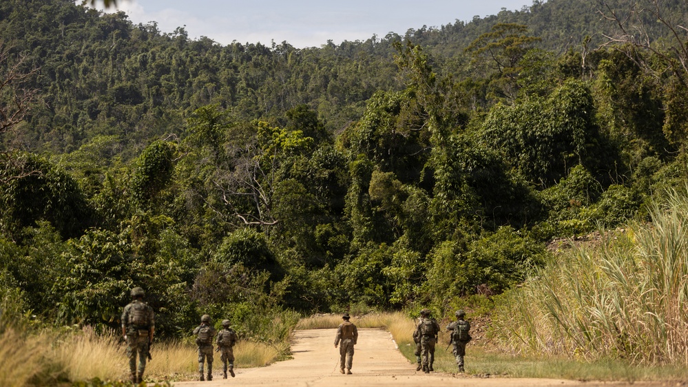 A group of approximately five armed military or security personnel are walking along a dirt road leading into dense tropical jungle. The soldiers are wearing camouflage uniforms and appear to be on patrol. The surrounding vegetation is lush and green, consistent with a tropical rainforest environment.