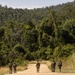 A group of approximately five armed military or security personnel are walking along a dirt road leading into dense tropical jungle. The soldiers are wearing camouflage uniforms and appear to be on patrol. The surrounding vegetation is lush and green, consistent with a tropical rainforest environment.