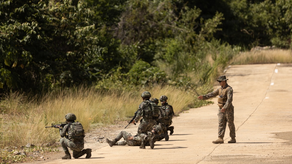 The image shows a military training exercise on a paved road bordered by dense vegetation. Several soldiers in camouflage uniforms are in tactical positions, with one individual appearing to act as an instructor or observer directing the others. The scene depicts a simulated combat or patrol scenario with soldiers carrying rifles and tactical gear.