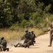 The image shows a military training exercise on a paved road bordered by dense vegetation. Several soldiers in camouflage uniforms are in tactical positions, with one individual appearing to act as an instructor or observer directing the others. The scene depicts a simulated combat or patrol scenario with soldiers carrying rifles and tactical gear.