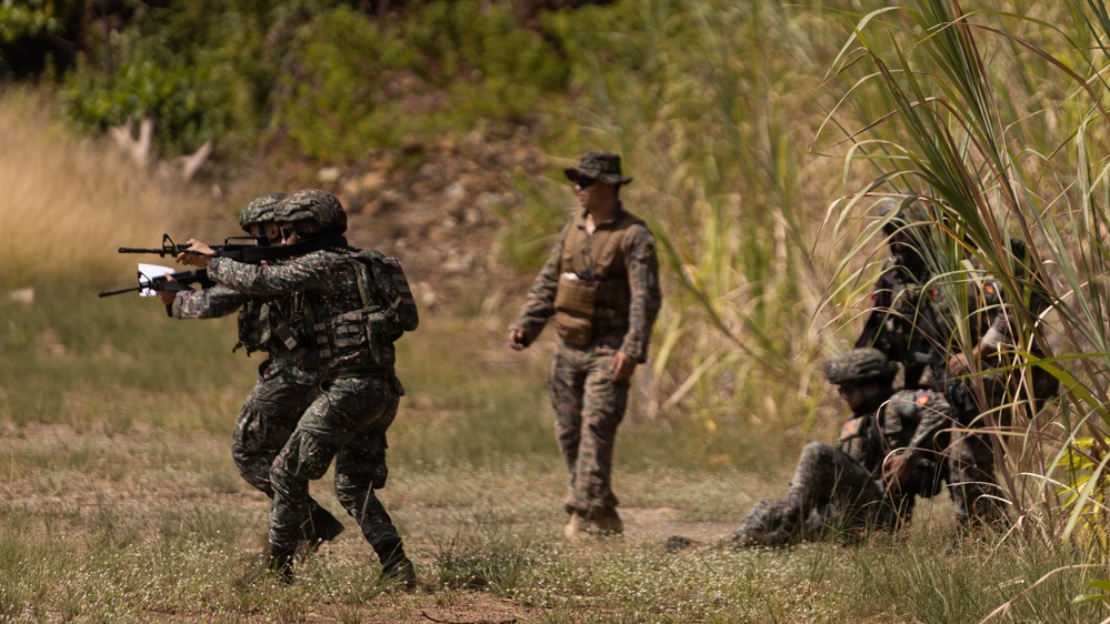 The image depicts a military training exercise in an outdoor, vegetated environment. Multiple soldiers in camouflage uniforms are engaged in tactical maneuvers, with one soldier in the foreground aiming a rifle while others take covered positions near tall grass. A supervising figure in a boonie hat and tactical vest observes the exercise from the center.