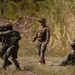 The image depicts a military training exercise in an outdoor, vegetated environment. Multiple soldiers in camouflage uniforms are engaged in tactical maneuvers, with one soldier in the foreground aiming a rifle while others take covered positions near tall grass. A supervising figure in a boonie hat and tactical vest observes the exercise from the center.