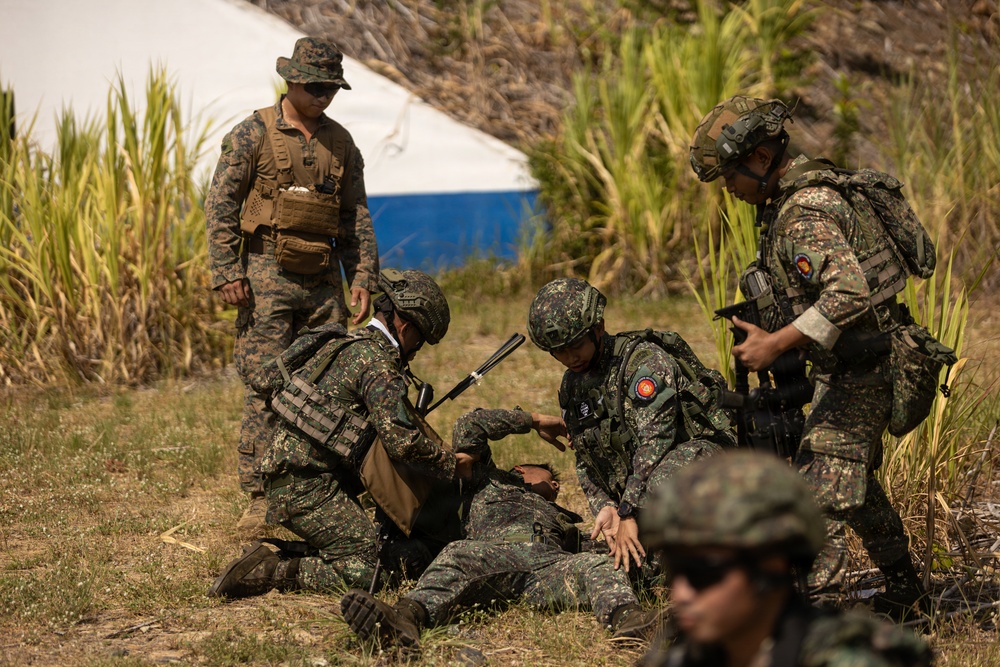 The image shows a group of military personnel in a field setting, appearing to conduct a combat casualty care or medical training exercise. One soldier is on the ground while others attend to them, suggesting a simulated casualty scenario. A soldier in US Marine Corps MARPAT camouflage observes the exercise alongside personnel in a different camouflage pattern, indicating a joint or combined training operation.