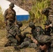 The image shows a group of military personnel in a field setting, appearing to conduct a combat casualty care or medical training exercise. One soldier is on the ground while others attend to them, suggesting a simulated casualty scenario. A soldier in US Marine Corps MARPAT camouflage observes the exercise alongside personnel in a different camouflage pattern, indicating a joint or combined training operation.