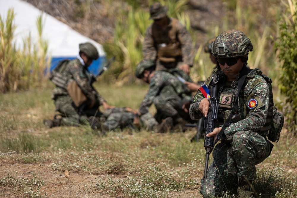 The image shows military personnel in camouflage uniforms conducting what appears to be a tactical training exercise or field operation. The foreground soldier is armed and in a kneeling security position, while personnel in the background appear to be performing casualty care or medical treatment on a downed individual. A Philippine flag patch is visible on the foreground soldier's uniform, identifying them as members of the Philippine Armed Forces.