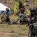 The image shows military personnel in camouflage uniforms conducting what appears to be a tactical training exercise or field operation. The foreground soldier is armed and in a kneeling security position, while personnel in the background appear to be performing casualty care or medical treatment on a downed individual. A Philippine flag patch is visible on the foreground soldier's uniform, identifying them as members of the Philippine Armed Forces.