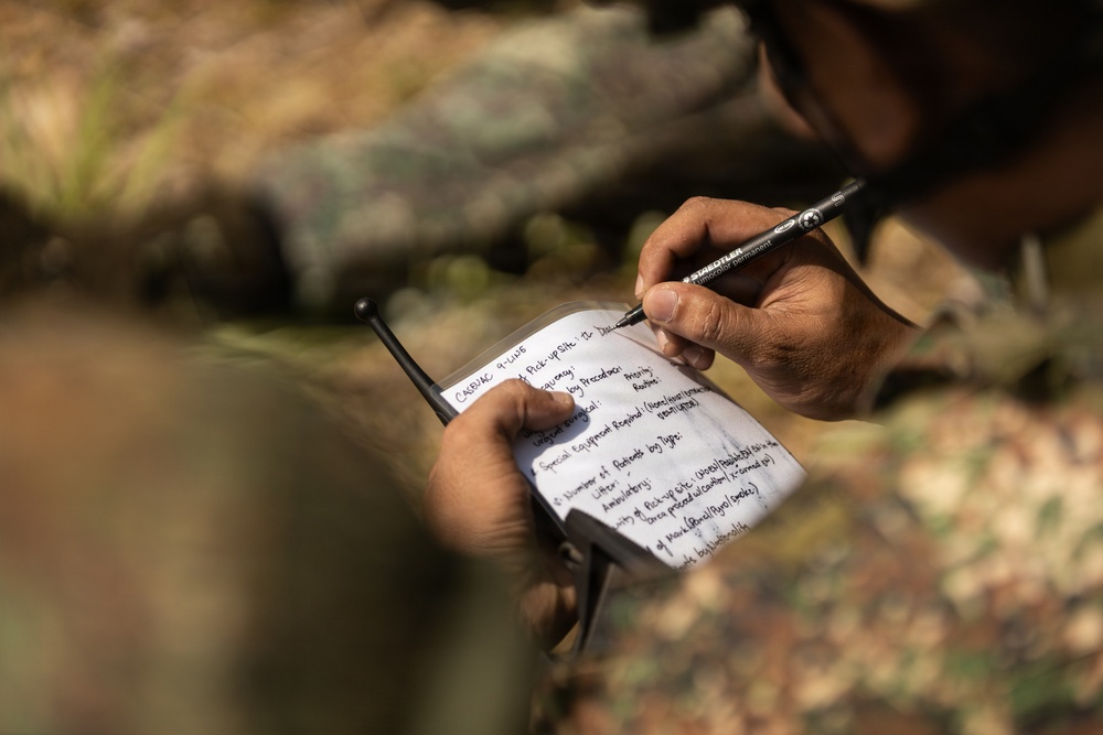 A person in camouflage military uniform is writing on a small notepad in an outdoor, wooded environment. The notepad contains a handwritten military CASEVAC (Casualty Evacuation) 9-Line report format, a standardized medical evacuation request used in military operations. The person is using a Staedtler Lumocolor permanent marker/pen to fill in the form.